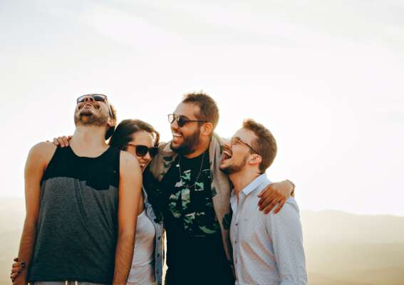 Three men and one woman standing together hugging each other in the outdoors with a light background and the sun shining