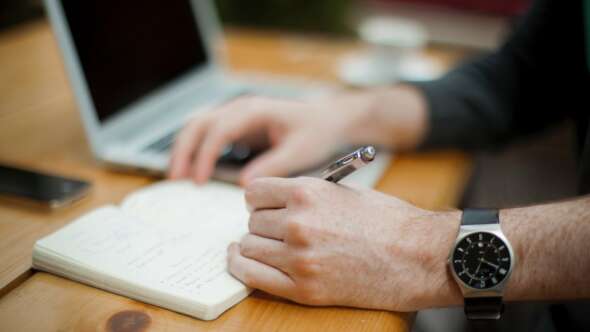 person writing in small journal on a bright wooden table with laptop in the background