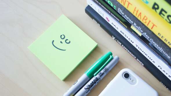 Light green post it note with a smiley face sitting on a desk with two sharpies and books