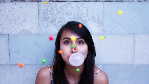 Young girl blowing a bubblegum bubble with skittles falling in front of camera shot