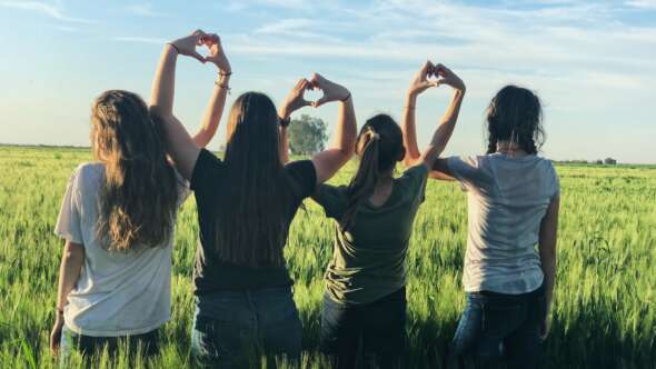 Four teen girls facing into a grassy field with their hands making heart shapes