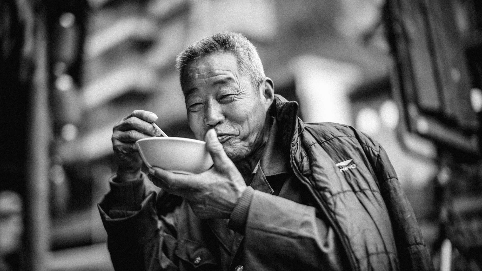 Monochrome portrait of older man eating from bowl with a cheeky grin on his face