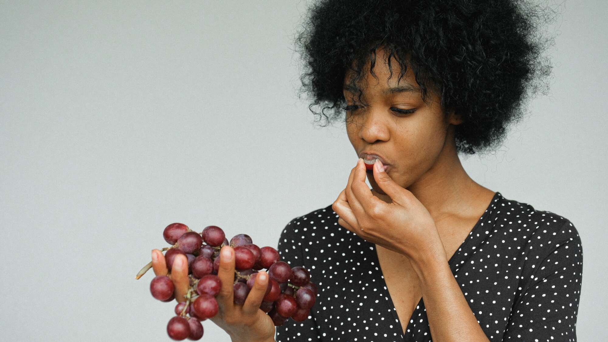 Photo of woman with black curly hair eating a handful of red grapes