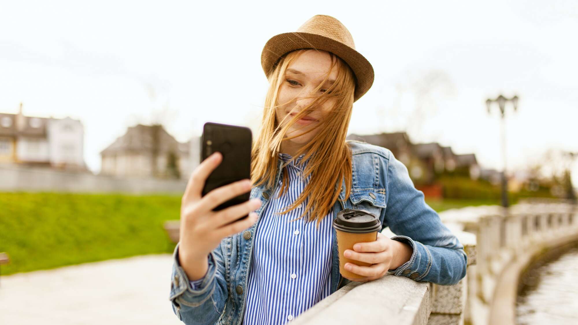 Woman waeing fedora and holding phone taking a selfie with coffee cup