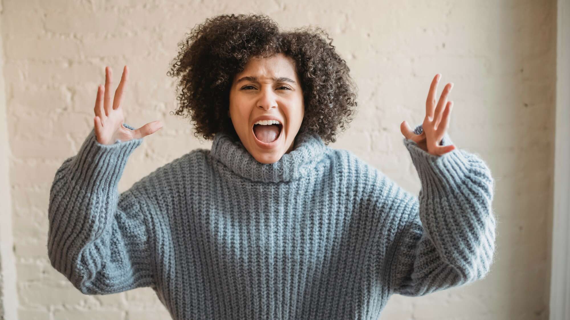 Woman with brown curly hair with hands in the air and her mouth open wide screaming