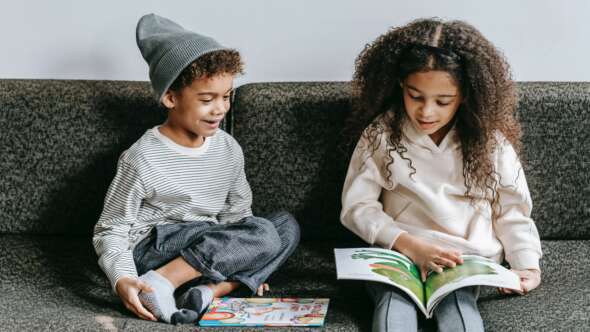 Young boy and girl sitting side by side on grey couch reading a cartoon book about a frog