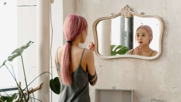 Girl with pink hair looking into mirror and putting lip gloss on lips with plant in the background