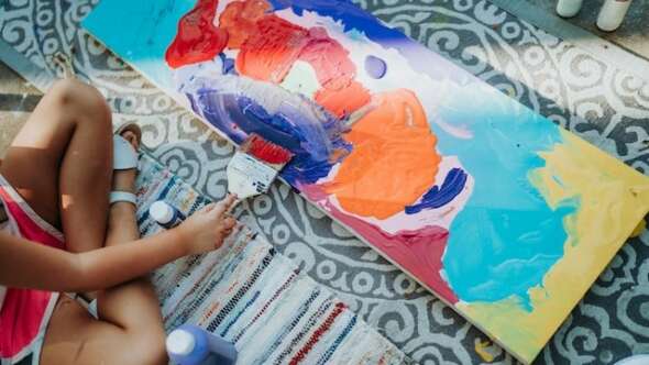 Child using large brush to paint on canvas yellow, blue, red and orange paints