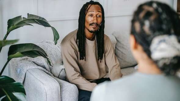 Man sitting on a grey couch next to a green leafy plant speaking to a female across from him