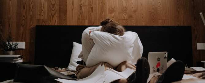 Woman on bed with pillow covering face with laptop and paperwork covering bed