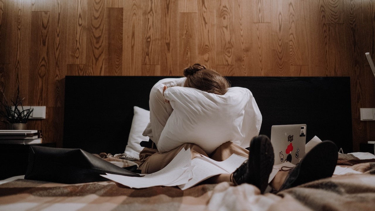Woman on bed with pillow covering face with laptop and paperwork covering bed