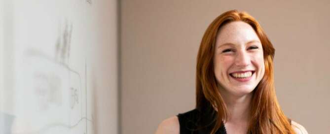 School teacher with red hair standing at whiteboard teaching.